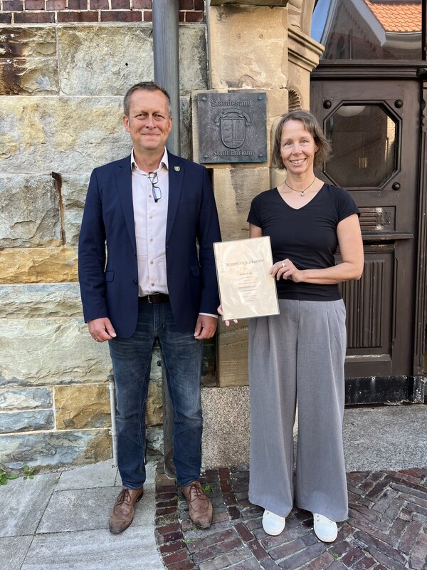 Bild vergr&ouml;&szlig;ern: B�rgermeister J�rgen Akkermann und Standesbeamtin Stefanie Byl stehen nebeneinander vor dem Rathaus. Frau Byl pr�sentiert ihre Ernennungsurkunde.